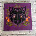 A framed artwork featuring a stylized black cat with flowers and leaves in a purple, green, and orange color scheme.