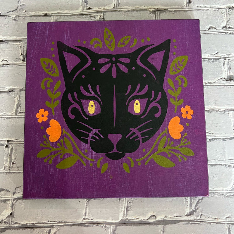 A framed artwork featuring a stylized black cat with flowers and leaves in a purple, green, and orange color scheme.