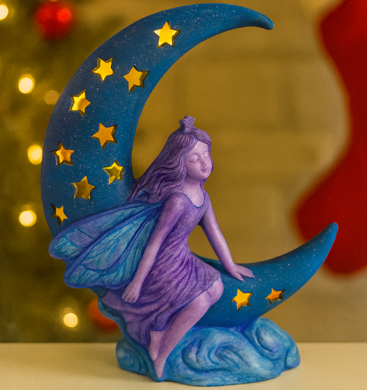 Decorative figurine of a fairy sitting on a blue crescent moon with stars, against a festive background.