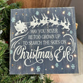 A lighted sign with a Christmas Eve theme, featuring a reindeer, snowflakes, and text on a wooden background, displayed on a Christmas tree.