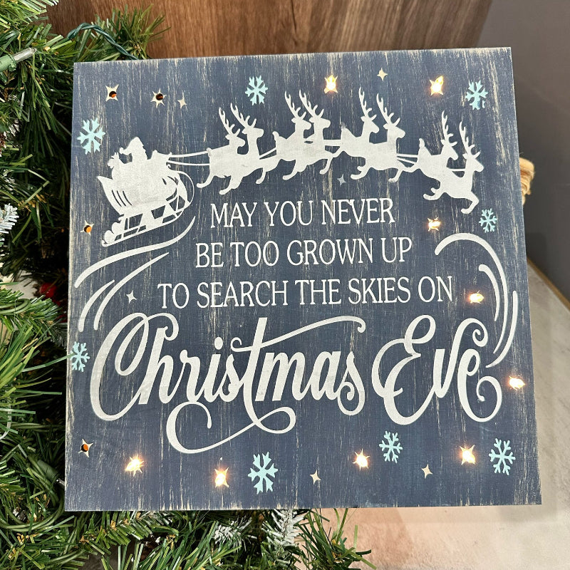 A lighted sign with a Christmas Eve theme, featuring a reindeer, snowflakes, and text on a wooden background, displayed on a Christmas tree.