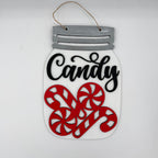 A decorative hanging sign shaped like a mason jar with a gray faux-metal lid, a hanging twine loop, a white body, black script text reading 'Candy,' and three red candy cane cutouts on a plain light background.