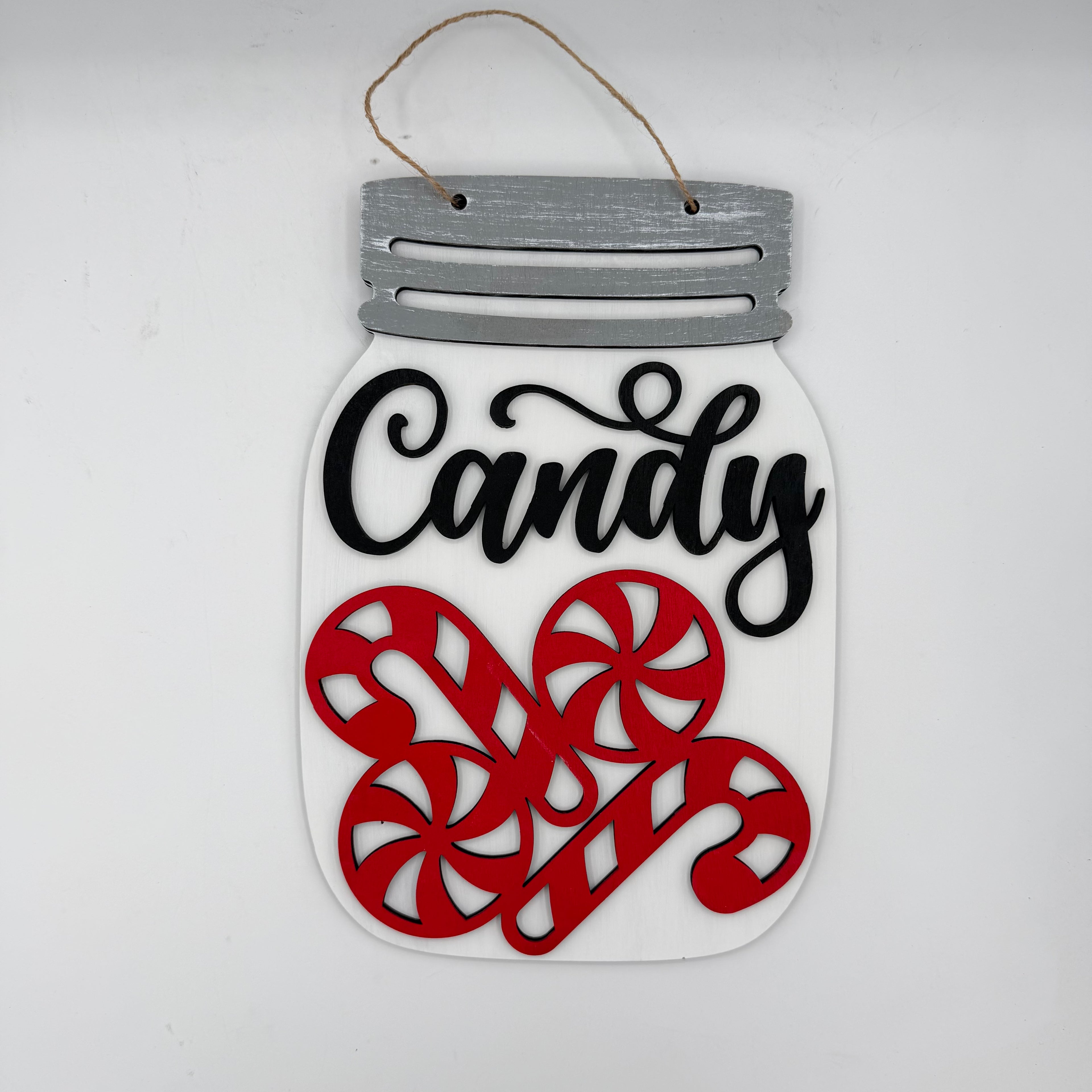 A decorative hanging sign shaped like a mason jar with a gray faux-metal lid, a hanging twine loop, a white body, black script text reading 'Candy,' and three red candy cane cutouts on a plain light background.