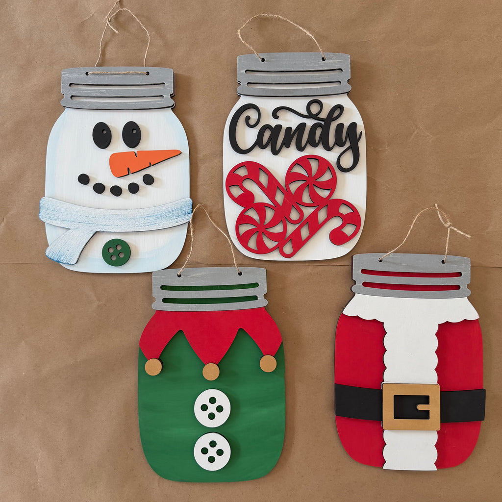 Four festive mason-jar-shaped hanging ornaments displayed on a brown kraft paper background. The ornaments are decorated with felt and paper crafts: a snowman with a light blue scarf, a candy jar with red peppermint candies and the word 'Candy' in black script, an elf with a green body, red collar, and white buttons, and a Santa Claus figure with a red and white body, black belt, and gold buckle.
