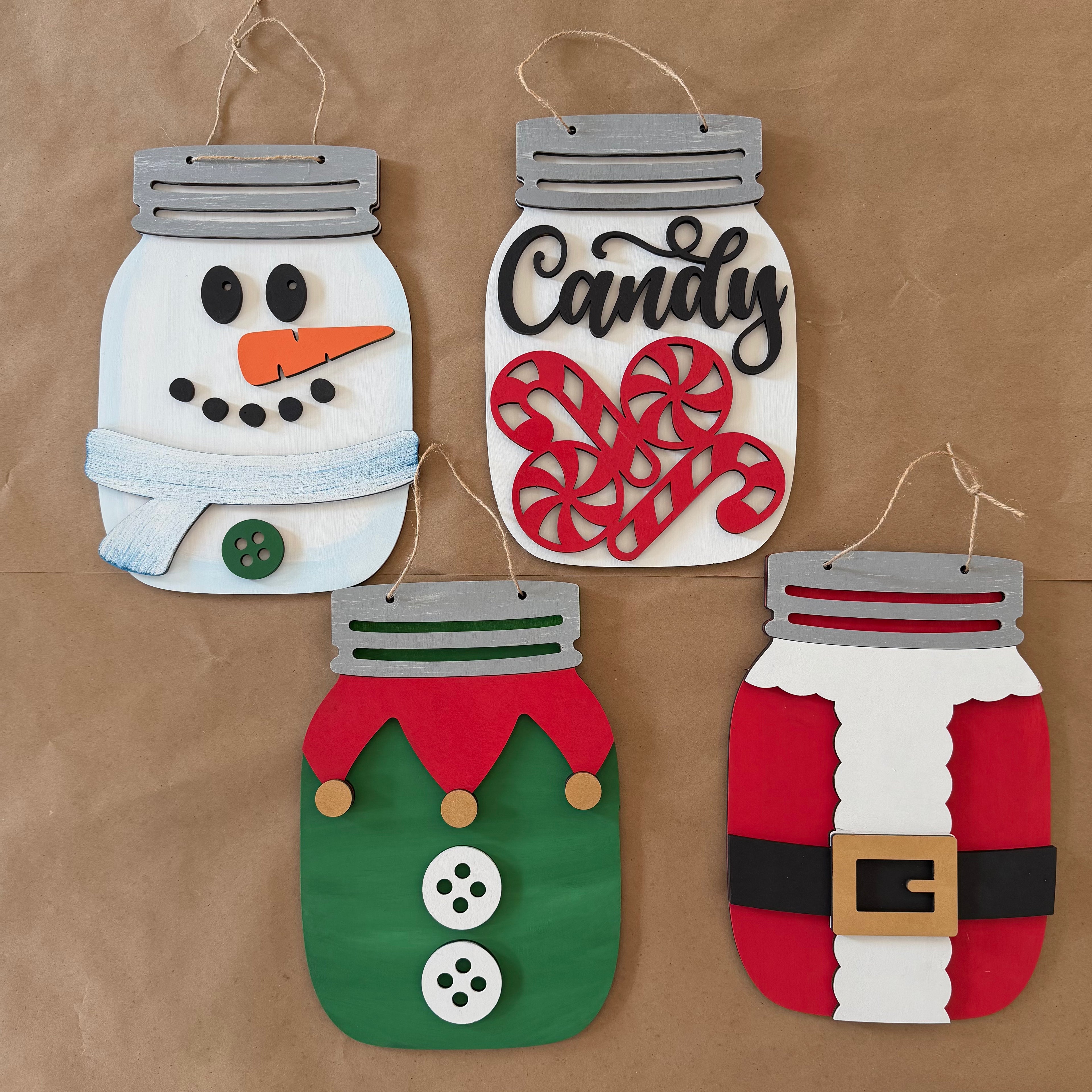 Four festive mason-jar-shaped hanging ornaments displayed on a brown kraft paper background. The ornaments are decorated with felt and paper crafts: a snowman with a light blue scarf, a candy jar with red peppermint candies and the word 'Candy' in black script, an elf with a green body, red collar, and white buttons, and a Santa Claus figure with a red and white body, black belt, and gold buckle.