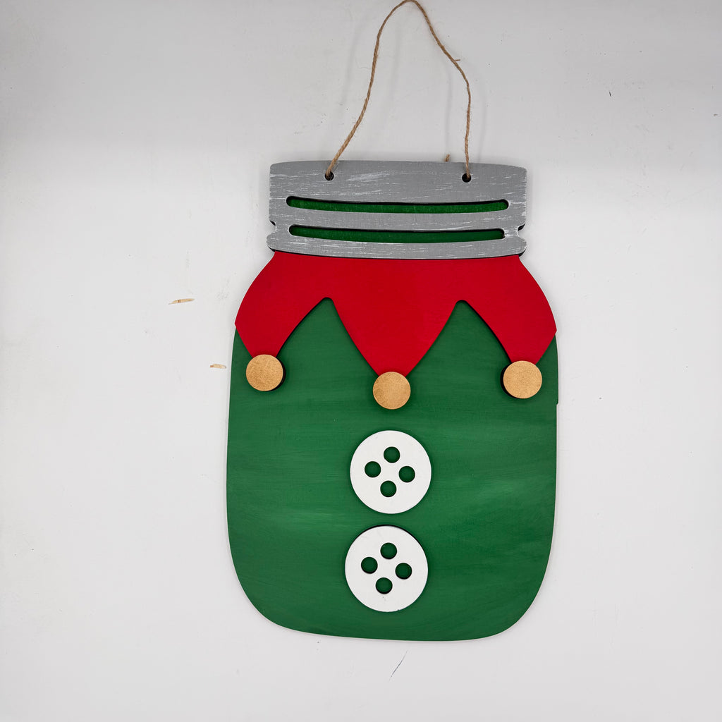 A handmade Christmas hanging ornament shaped like a mason jar with a green body, red hat, white buttons, and a silver lid, suspended by a twine string.