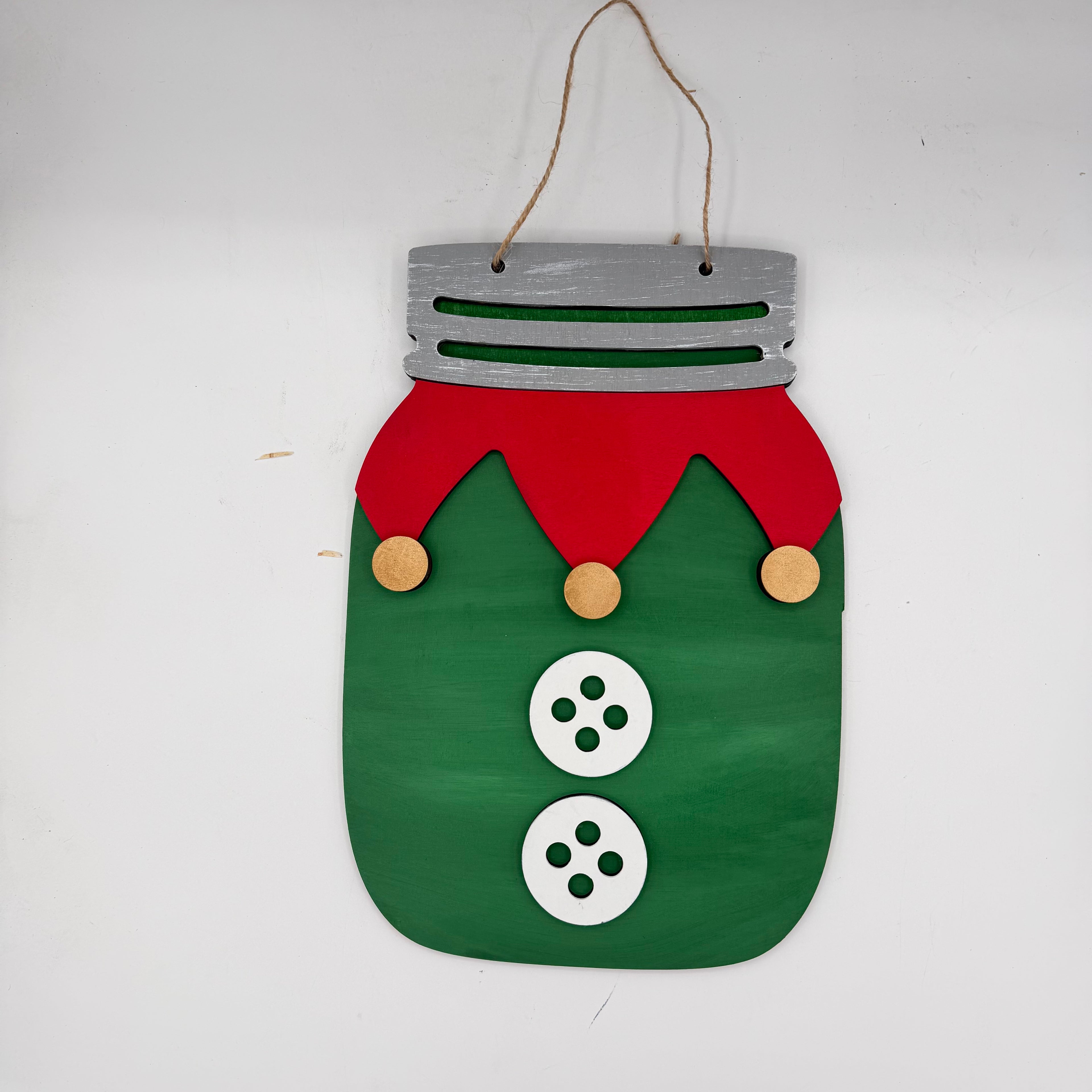 A handmade Christmas hanging ornament shaped like a mason jar with a green body, red hat, white buttons, and a silver lid, suspended by a twine string.