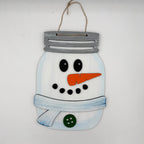 A decorative wooden snowman hanging ornament designed like a mason jar, with a gray painted lid, white body, black facial features, an orange carrot nose, a blue scarf, and a green button, hanging against a plain white wall.
