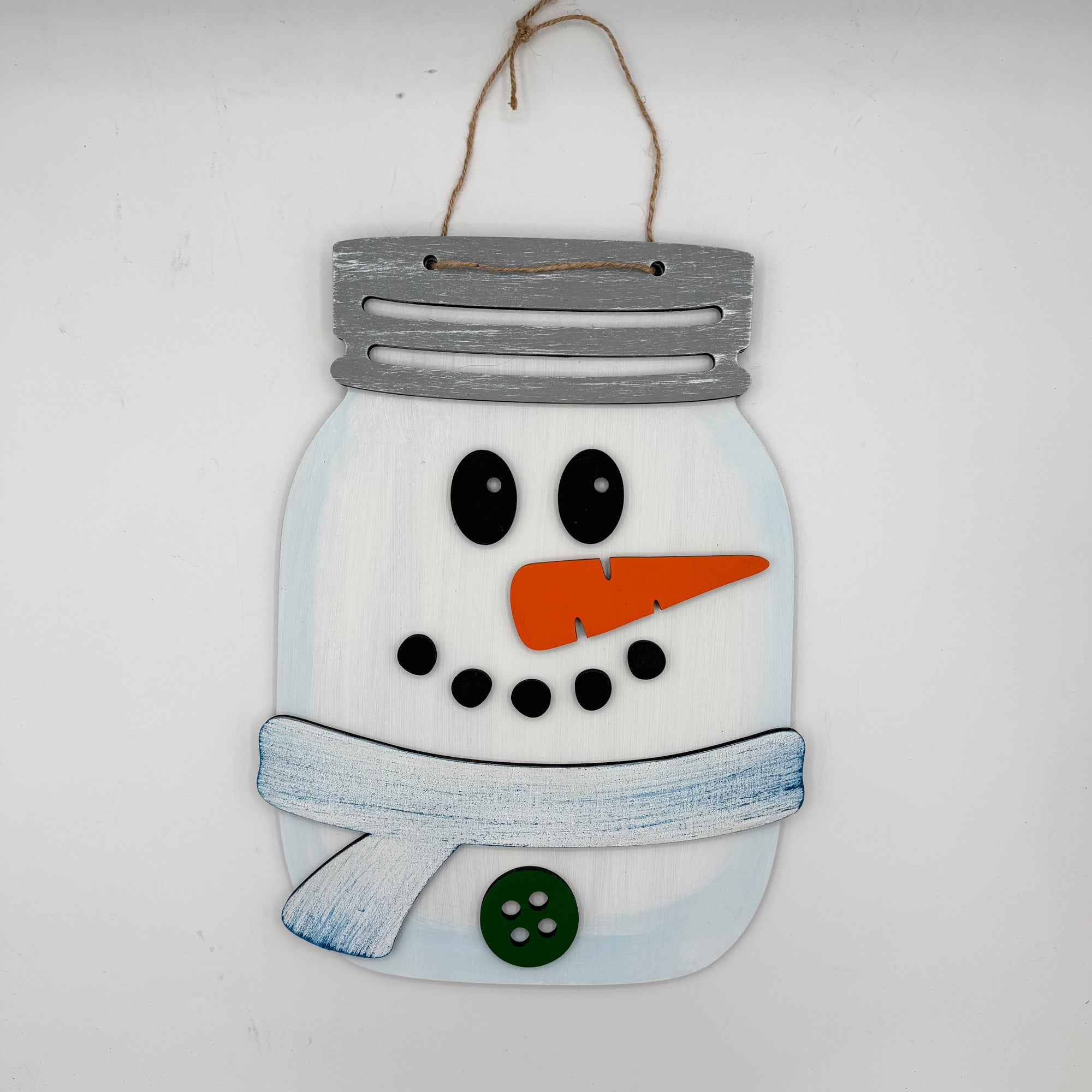 A decorative wooden snowman hanging ornament designed like a mason jar, with a gray painted lid, white body, black facial features, an orange carrot nose, a blue scarf, and a green button, hanging against a plain white wall.