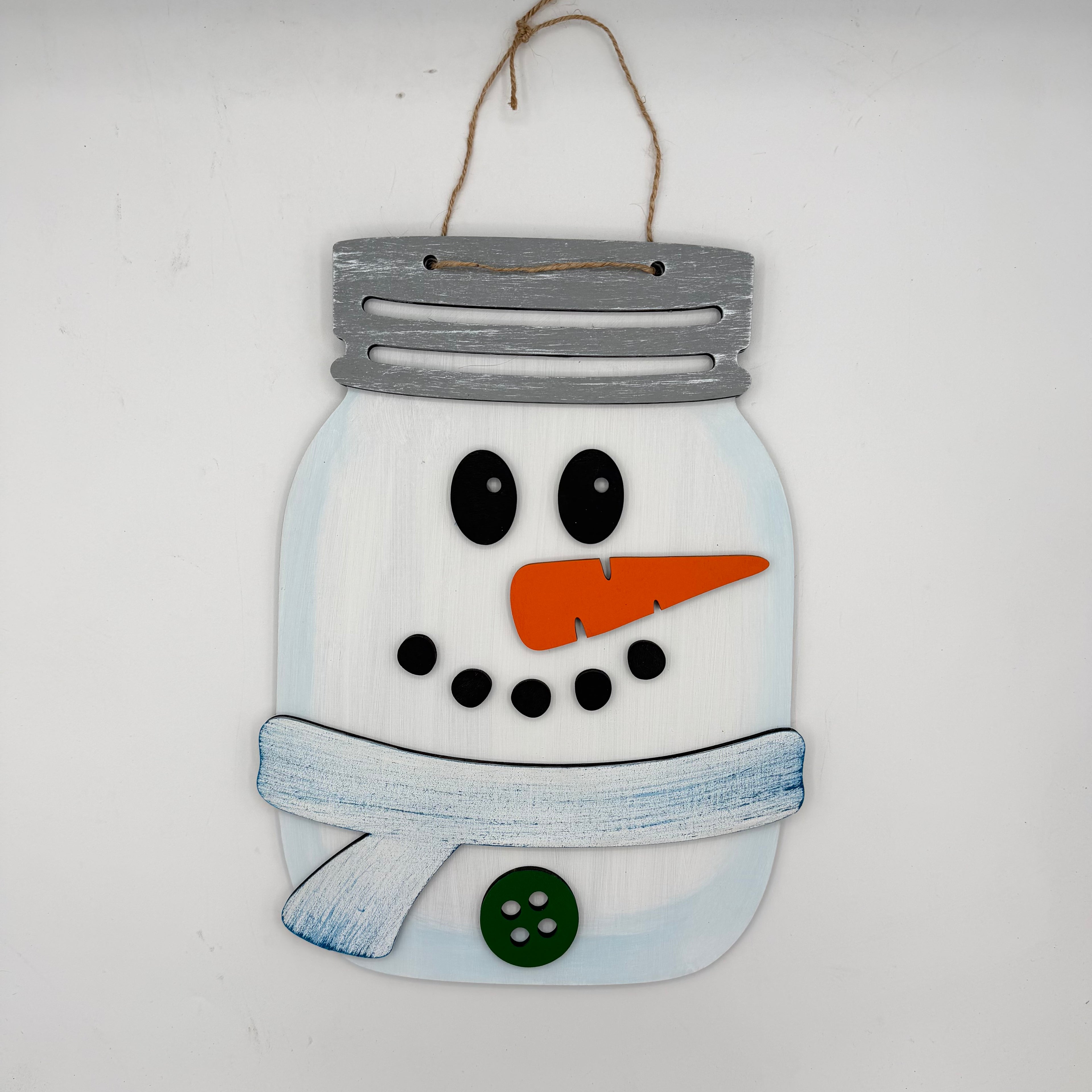 A decorative wooden snowman hanging ornament designed like a mason jar, with a gray painted lid, white body, black facial features, an orange carrot nose, a blue scarf, and a green button, hanging against a plain white wall.