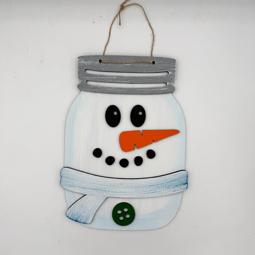 A decorative wooden snowman hanging ornament designed like a mason jar, with a gray painted lid, white body, black facial features, an orange carrot nose, a blue scarf, and a green button, hanging against a plain white wall.