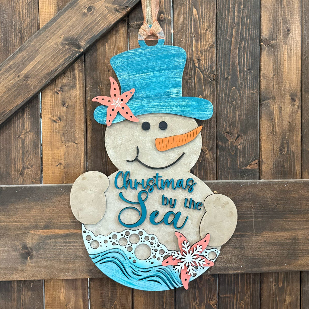 A hand-painted wooden Christmas snowman hanging on a rustic dark wood plank wall. The snowman features a blue top hat decorated with a coral starfish, a blue carrot nose, and the text 'Christmas by the Sea' painted on its belly with blue waves at the bottom.