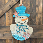A hand-painted wooden Christmas snowman hanging on a rustic dark wood plank wall. The snowman features a blue top hat decorated with a coral starfish, a blue carrot nose, and the text 'Christmas by the Sea' painted on its belly with blue waves at the bottom.