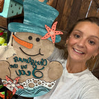 A smiling woman holds up a large decorative holiday sign shaped like a snowman in a beach theme. The sign features a teal top hat, orange starfish accents, a carrot nose, and the words 'Christmas on the Sea' in teal script over a tan background.