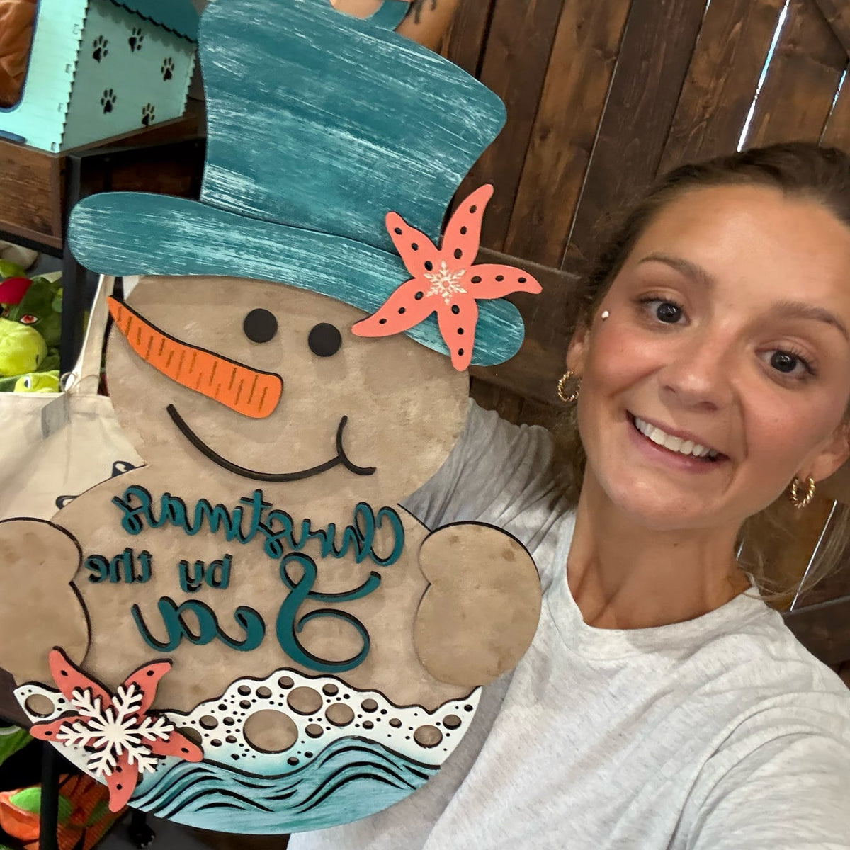 A smiling woman holds up a large decorative holiday sign shaped like a snowman in a beach theme. The sign features a teal top hat, orange starfish accents, a carrot nose, and the words 'Christmas on the Sea' in teal script over a tan background.