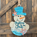 A hand-painted wooden Christmas snowman hanging on a rustic dark wood plank wall. The snowman features a blue top hat decorated with a coral starfish, a blue carrot nose, and the text 'Christmas by the Sea' painted on its belly with blue waves at the bottom.