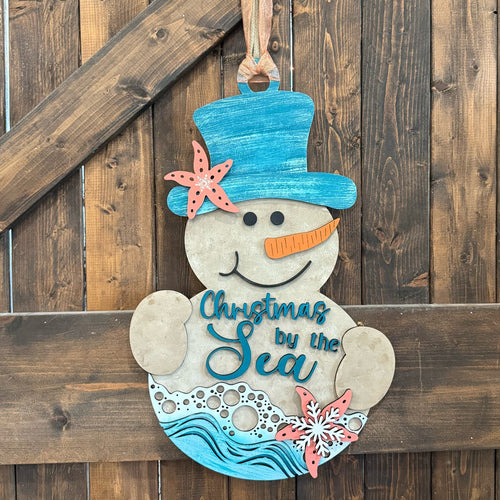 A hand-painted wooden Christmas snowman hanging on a rustic dark wood plank wall. The snowman features a blue top hat decorated with a coral starfish, a blue carrot nose, and the text 'Christmas by the Sea' painted on its belly with blue waves at the bottom.
