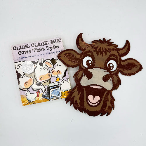 A cartoon cow head cutout or sticker placed next to a small children's book titled "Click, Clack, Moo Cows That Type" on a plain white background.
