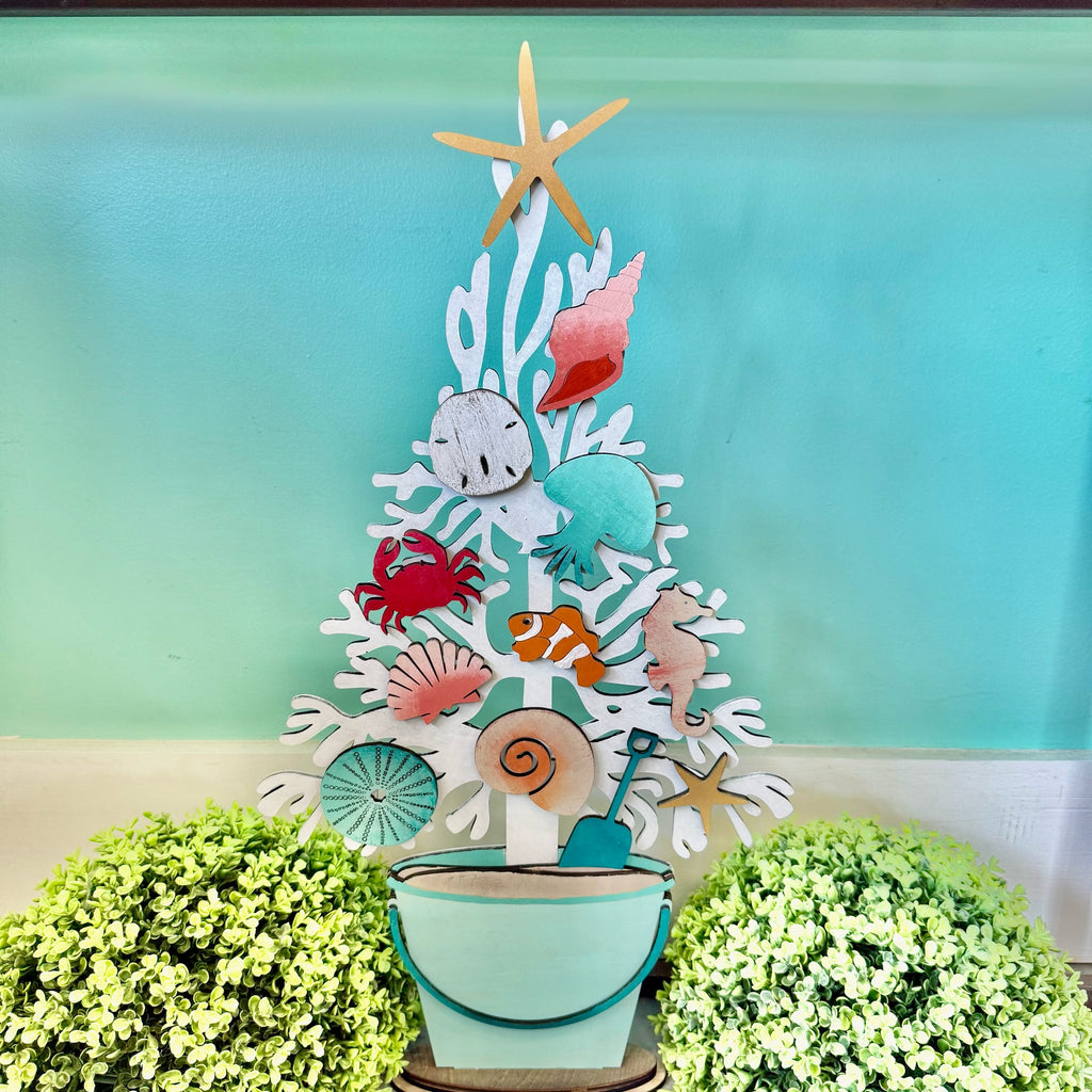 A decorative white coral tree centerpiece with colorful seashell and sea life cutouts, including a starfish, crab, fish, octopus, and seahorse, displayed in a small bucket with a shovel, set against a turquoise background with green foliage at the bottom.