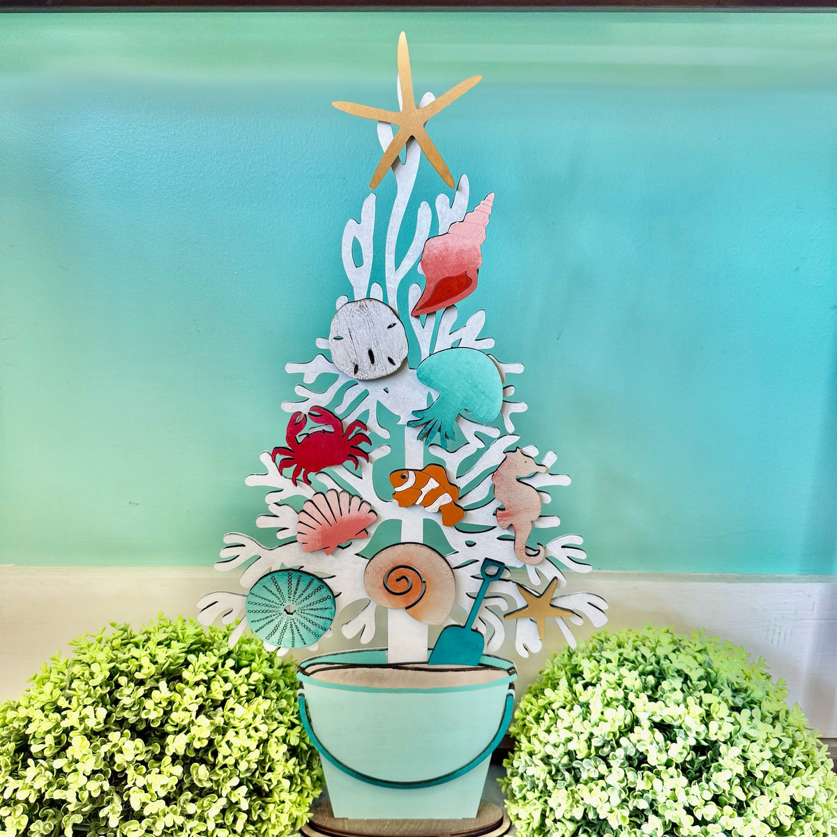 A decorative white coral tree centerpiece with colorful seashell and sea life cutouts, including a starfish, crab, fish, octopus, and seahorse, displayed in a small bucket with a shovel, set against a turquoise background with green foliage at the bottom.