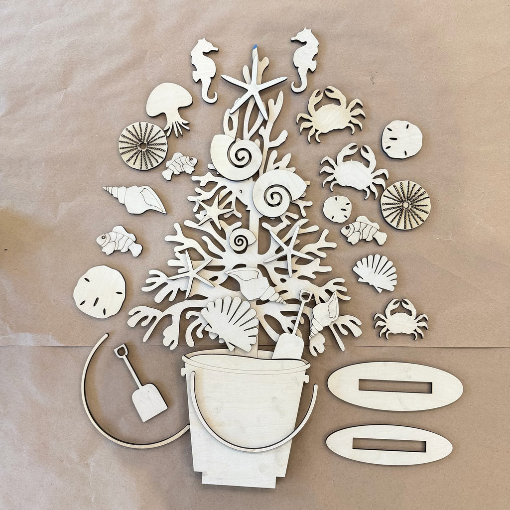 A flat-lay arrangement of light-colored wooden beach-themed cutouts and shapes, including a bucket, starfish, seahorse, crabs, shells, sand dollars, a shovel, and two oval pieces, laid on a tan background.