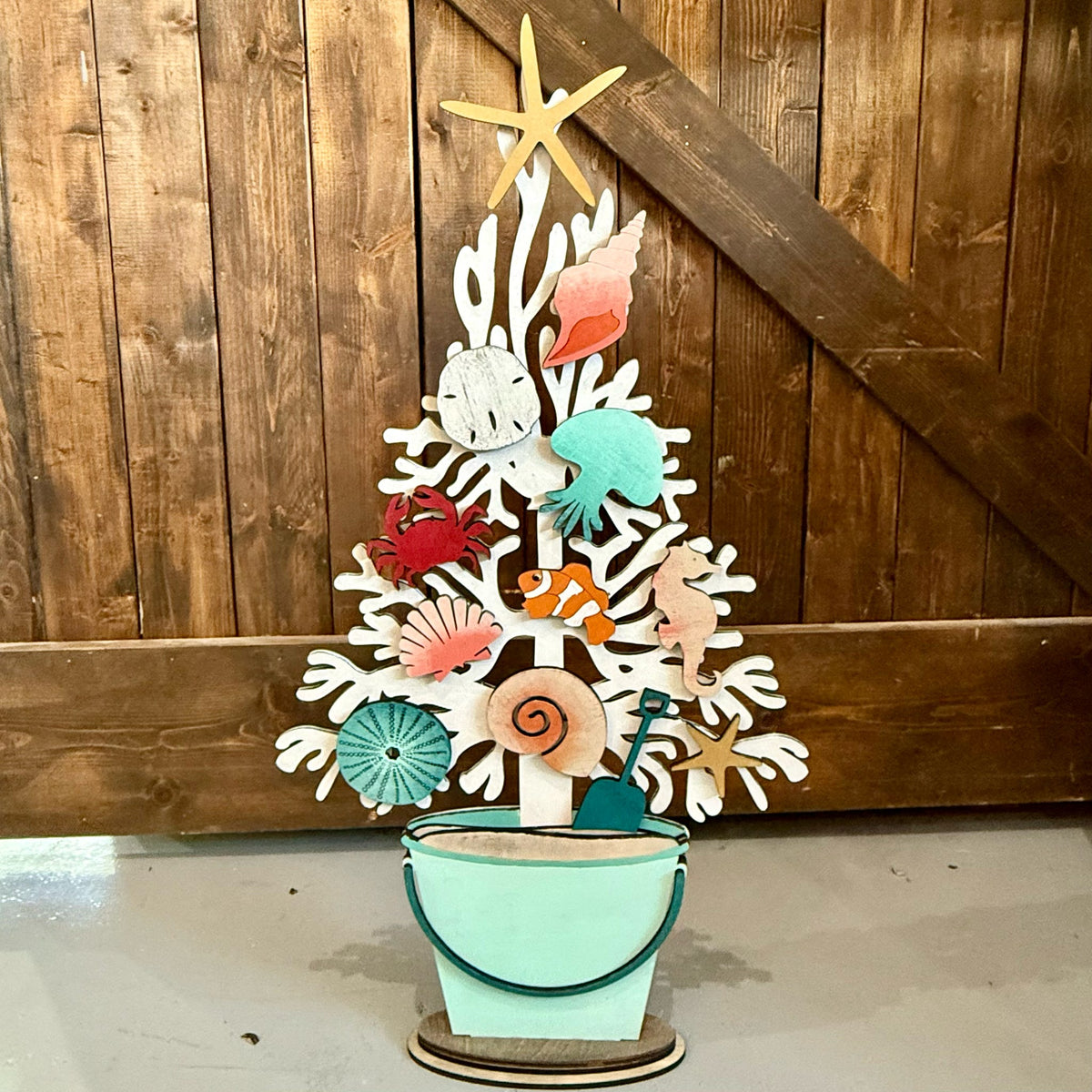 A decorative wooden beach-themed tree ornament with colorful seashells, starfish, fish, and a mint green bucket base, displayed against a rustic wooden plank wall.