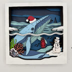 A square layered paper-cut style wall art piece showing a blue whale swimming underwater, with a red Santa hat, coral, a red lantern, and a snowman on the ocean floor. The artwork is mounted on a white board against a light gray background.