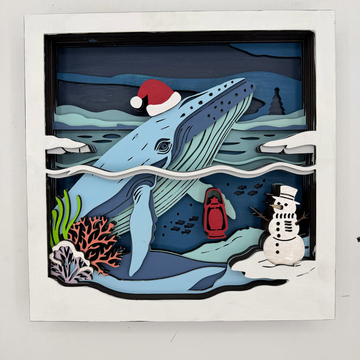 A square layered paper-cut style wall art piece showing a blue whale swimming underwater, with a red Santa hat, coral, a red lantern, and a snowman on the ocean floor. The artwork is mounted on a white board against a light gray background.