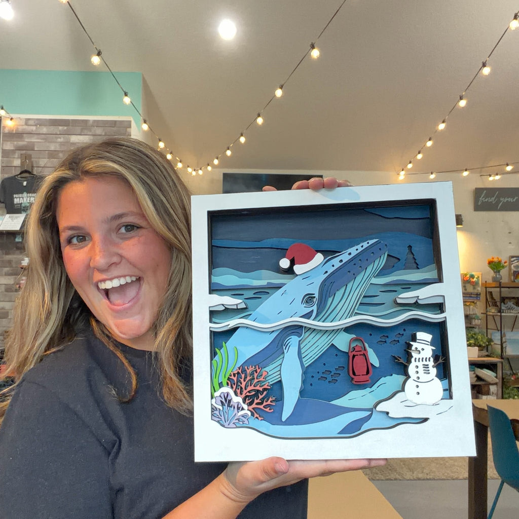 A woman smiling in a craft studio holds up a square layered paper art piece depicting an underwater winter scene with a blue whale, snowman, coral, and a Santa hat.