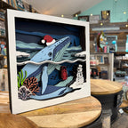 A square white-framed layered paper-cut wall art piece showing an underwater winter scene with a blue whale wearing a red Santa hat, a snowman, coral, and a red life vest, displayed on a wooden table in a shop.
