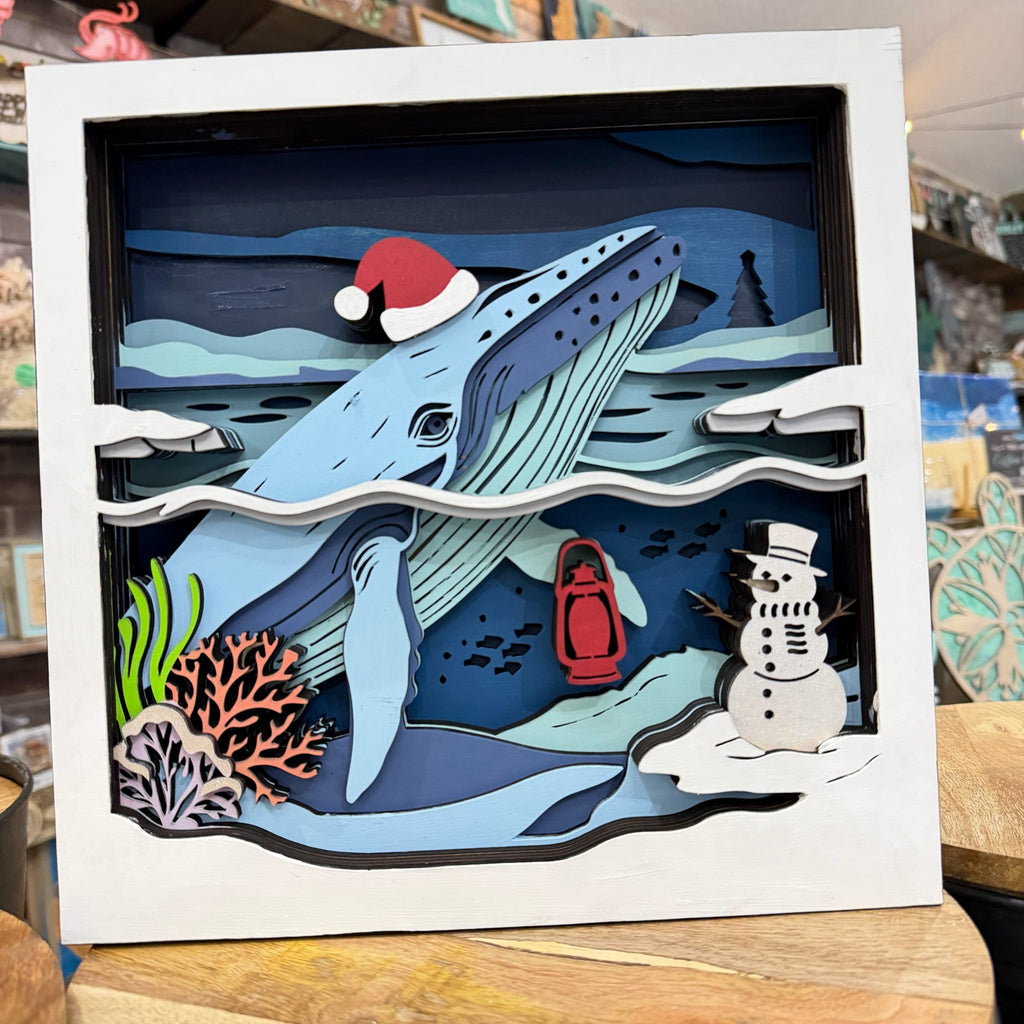 A square white shadow box artwork featuring a layered paper-cut winter ocean scene with a blue whale wearing a red Santa hat, a snowman, coral, and a red lantern, displayed on a wooden surface.