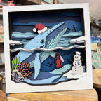A square white shadow box artwork featuring a layered paper-cut winter ocean scene with a blue whale wearing a red Santa hat, a snowman, coral, and a red lantern, displayed on a wooden surface.