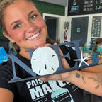 A smiling woman holds a navy blue wooden decorative sign spelling 'MEL' with a white sand dollar and starfish cutouts, in a workshop or craft space.