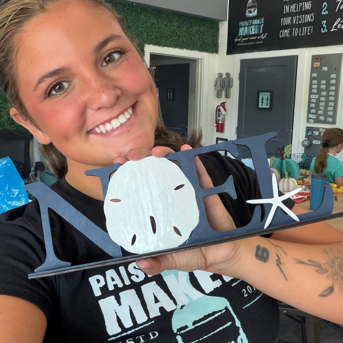 A smiling woman holds a navy blue wooden decorative sign spelling 'MEL' with a white sand dollar and starfish cutouts, in a workshop or craft space.