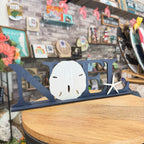 A navy blue wooden decorative sign spelling 'NOLA' sits on a round wooden table in a shop. The 'O' is shaped like a white sand shell, and there is a small white starfish on the 'A'.