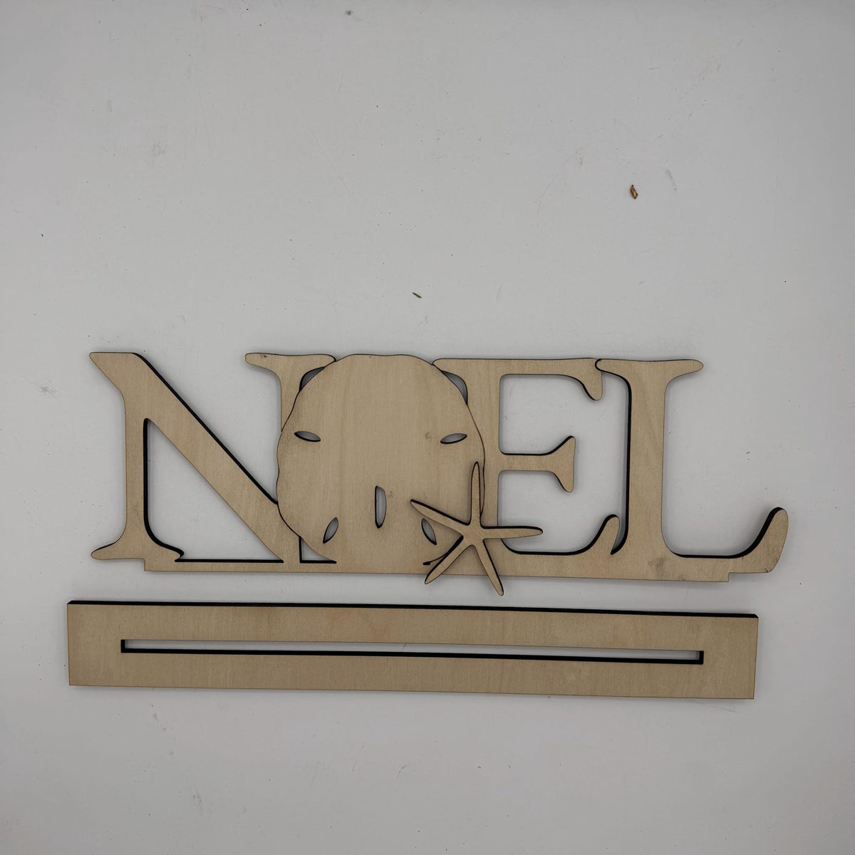 A wooden personalized name sign spelling 'NOEL' with a sand dollar and starfish cutout, mounted on a plain white wall above a long rectangular wooden base.