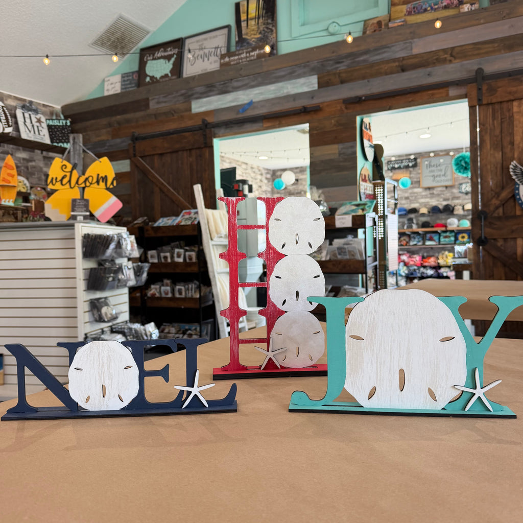 Three decorative wooden letters spelling NOEL, JOLY, and JOLY are displayed on a tabletop inside a rustic gift shop. The letters feature distressed wood finishes in navy blue, red, and teal, with shell and starfish accents.