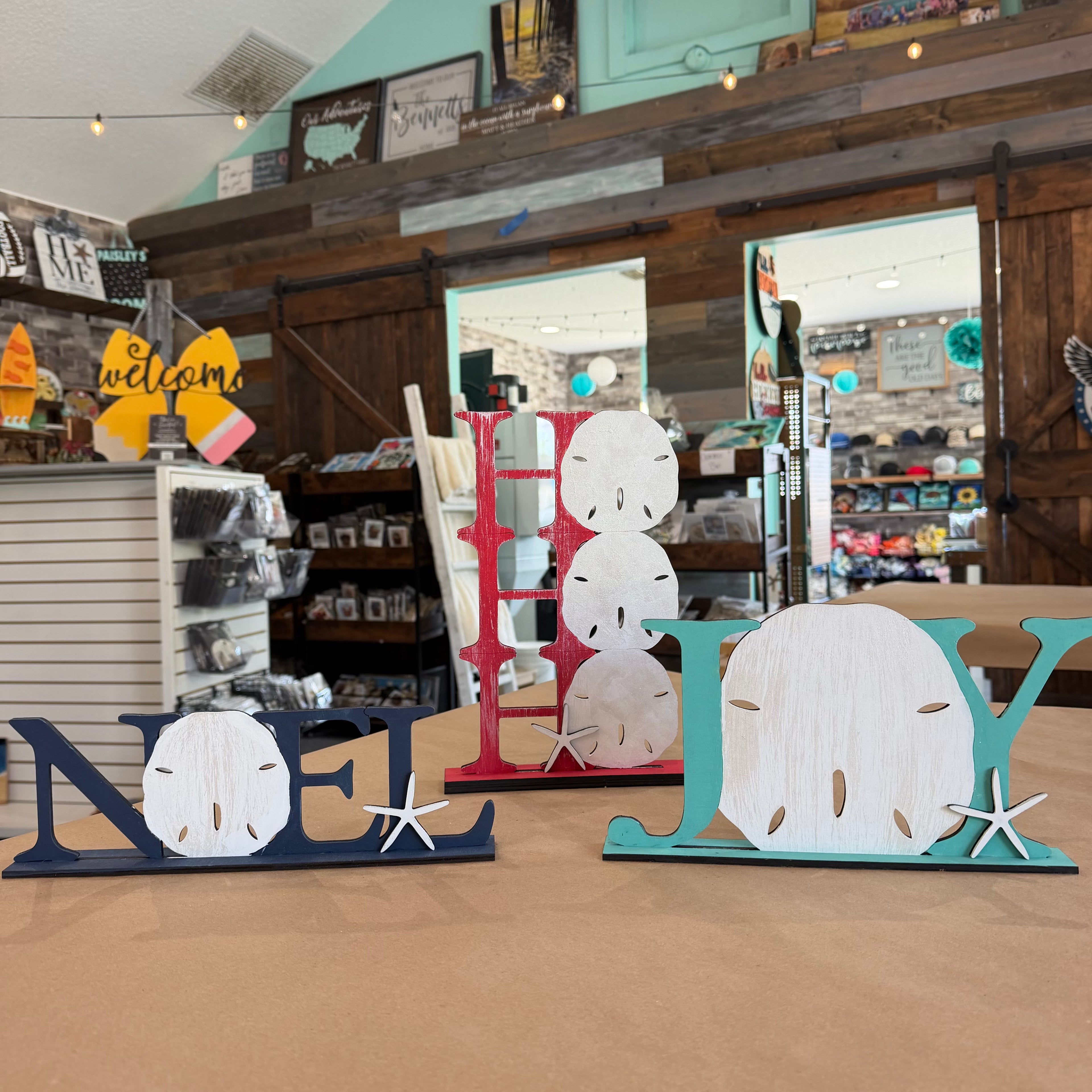 Three decorative wooden letters spelling NOEL, JOLY, and JOLY are displayed on a tabletop inside a rustic gift shop. The letters feature distressed wood finishes in navy blue, red, and teal, with shell and starfish accents.