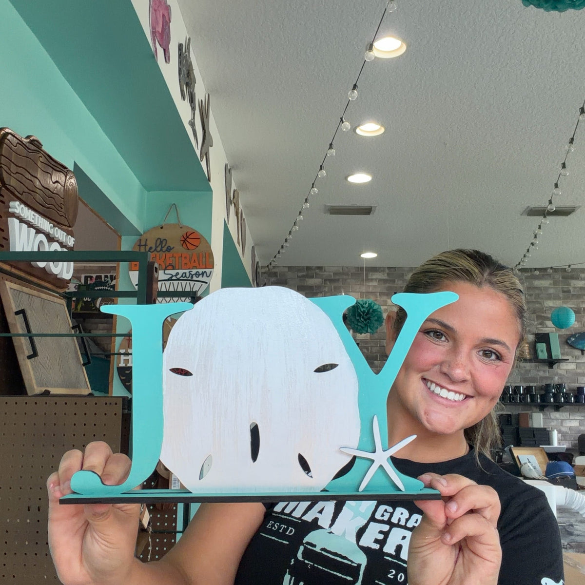 A smiling woman holds up a decorative wooden sign spelling 'JOY' with a white sand shell in the letter O, set against a turquoise background. The sign is photographed in a retail store setting.