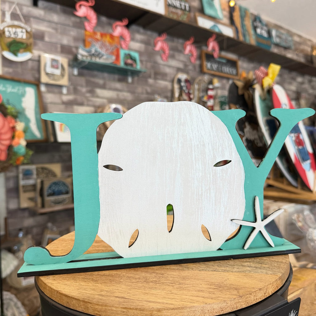 A decorative wooden sign spelling 'JOY' in turquoise and white, styled with a sand dollar in the center and a white starfish accent, displayed on a wooden pedestal in a shop setting.