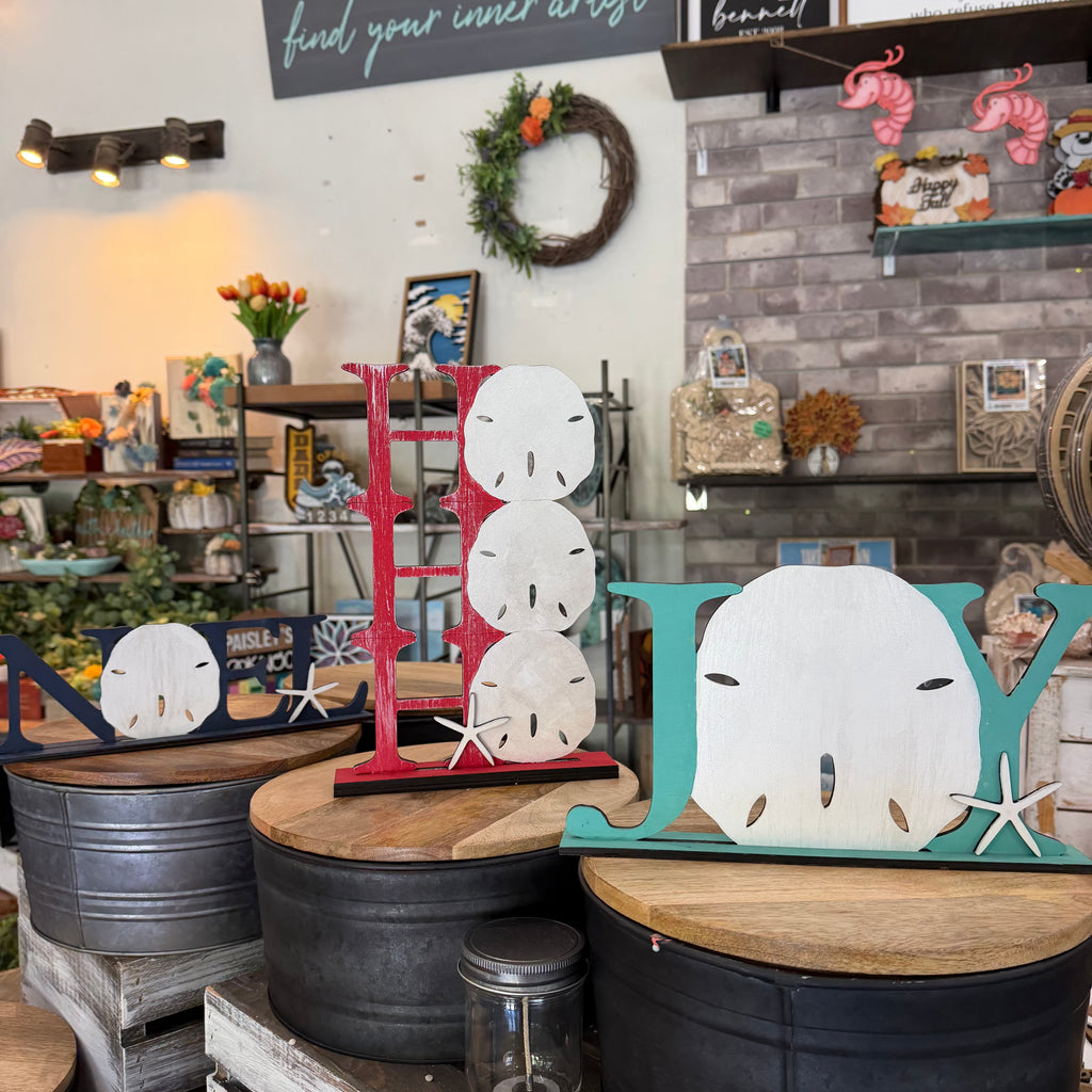 Decorative wooden letters spelling 'JAY' styled in a rustic shop setting, featuring a red ladder-style 'J', a teal sand dollar with starfish accent 'Y', and smaller matching sand dollar and starfish pieces on metal bins.