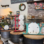 Decorative wooden letters spelling 'JAY' styled in a rustic shop setting, featuring a red ladder-style 'J', a teal sand dollar with starfish accent 'Y', and smaller matching sand dollar and starfish pieces on metal bins.