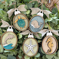 Five oval wooden hanging ornaments with burlap ribbon loops, each featuring a different sea-themed design: a turtle, a whale, a dolphin, a starfish, and a seahorse. The ornaments are arranged on green foliage.