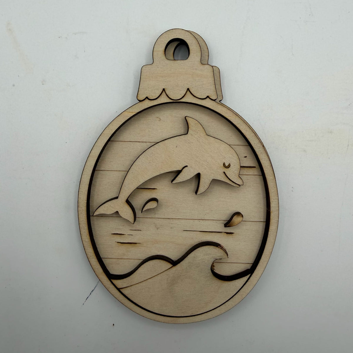 A laser-cut wooden ornament or plaque featuring a dolphin jumping over stylized waves, shown on a plain off-white background.
