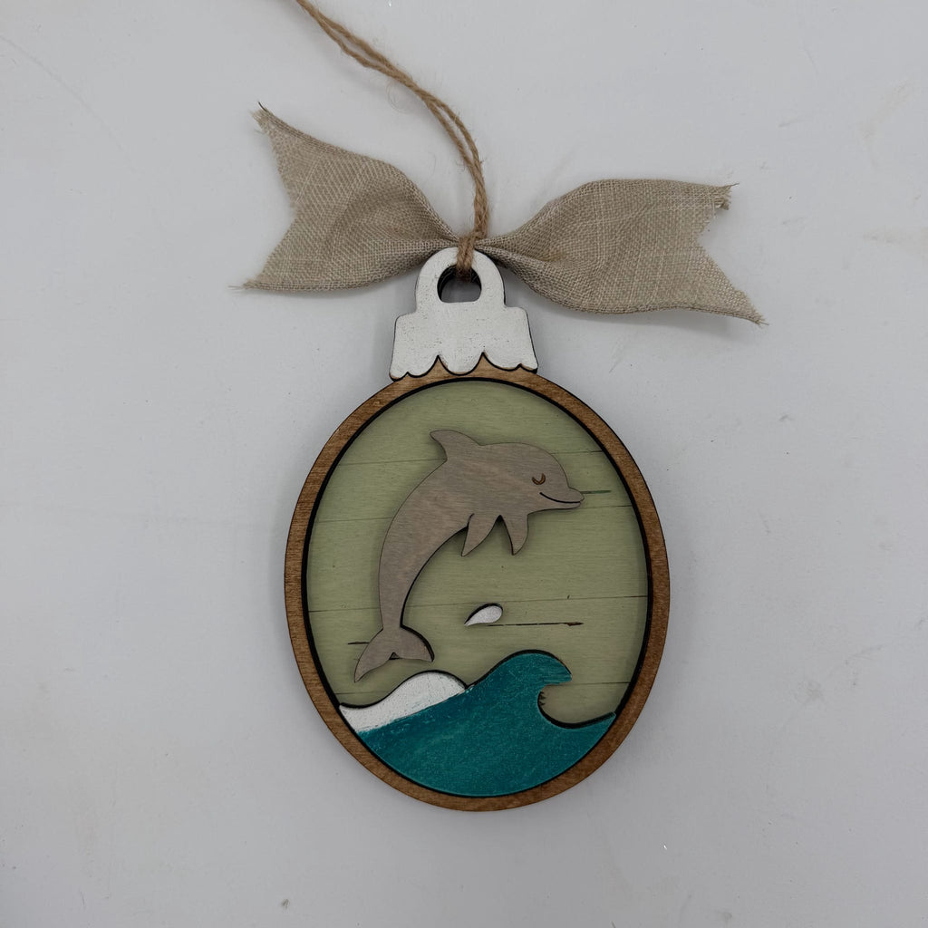 A handcrafted oval wooden ornament featuring a laser-cut design of a jumping dolphin over teal and white ocean waves, hanging from a rustic burlap ribbon bow on a light gray background.