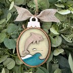 A round wooden hanging ornament featuring a layered design with a dolphin jumping over blue and white waves, tied with a burlap bow and displayed among green eucalyptus leaves.