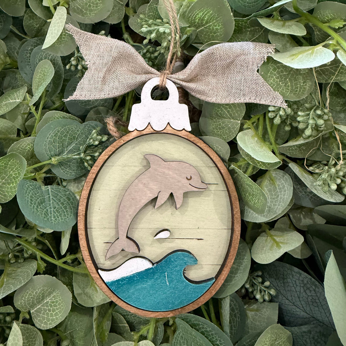 A round wooden hanging ornament featuring a layered design with a dolphin jumping over blue and white waves, tied with a burlap bow and displayed among green eucalyptus leaves.