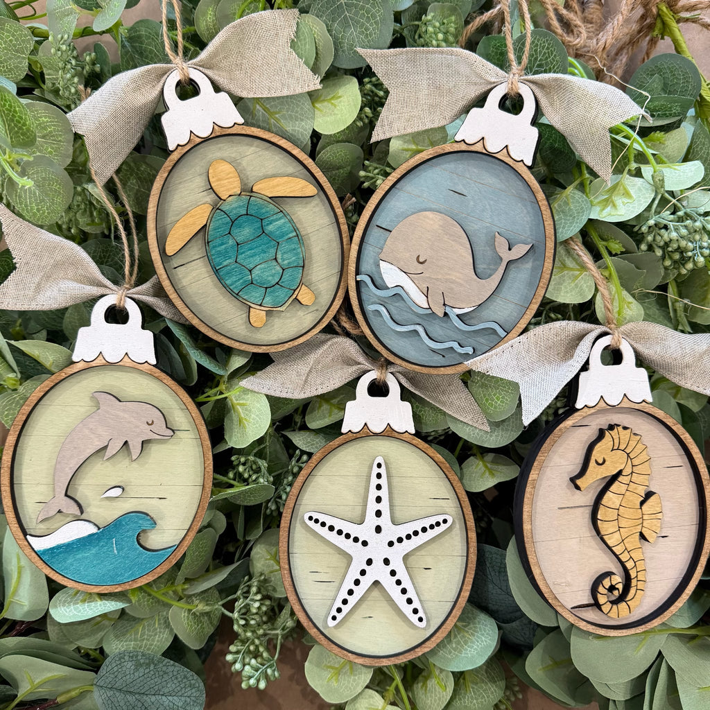 Six oval wooden hanging ornaments with burlap bows, featuring sea-themed designs including a turtle, whale, dolphin, starfish, and seahorse, displayed among green leaves.