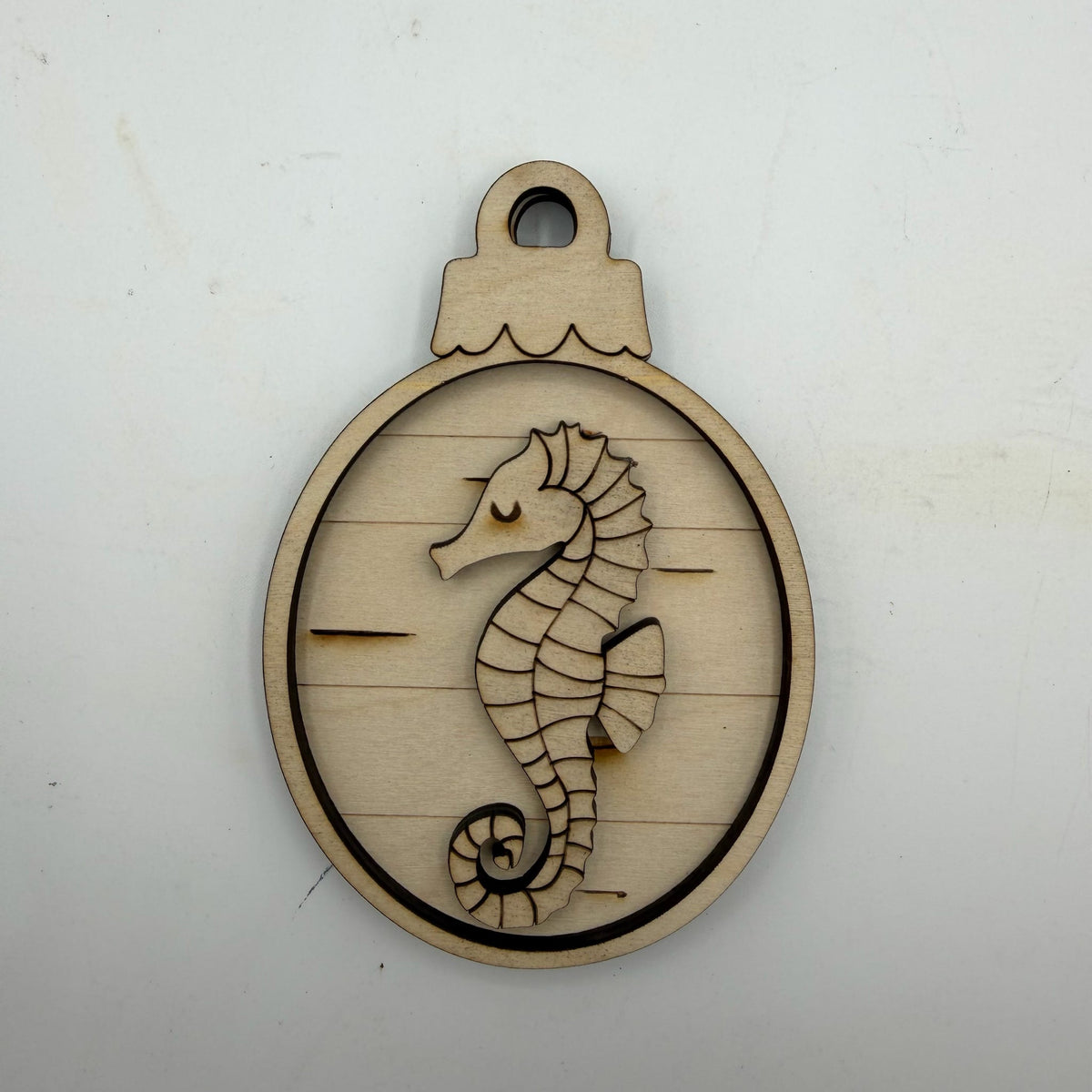A round laser-cut wooden ornament or pendant featuring a seahorse design on a light natural wood background, photographed against a plain off-white surface.