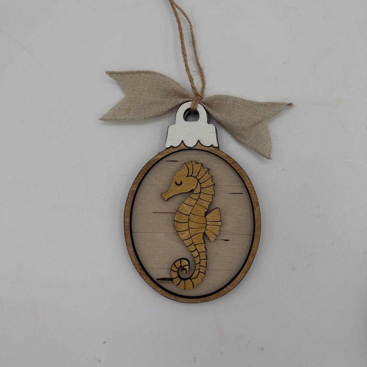 A wooden oval hanging ornament featuring a laser-cut seahorse design in the center, suspended from a twine string with a burlap ribbon bow, shown against a plain off-white background.
